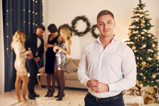 Man in formal clothes standing in front of his friends. Group of people have a new year party indoors together.