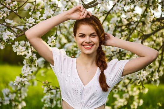 portrait of a joyful woman in a light dress against the background of a flowering tree, raising her hands to her head. High quality photo