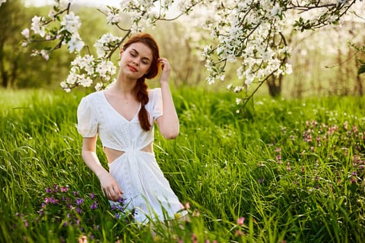 a young woman in a light summer dress stands near a tree with flowers and smiles looking at the camera. High quality photo
