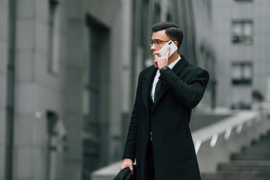 Talking by phone. Businessman in black suit and tie is outdoors in the city.