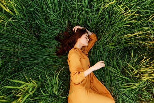 an elegant woman lies in the tall green grass in a long summer orange dress and holding her hair with her hands enjoys nature and sunny weather with her eyes closed. High quality photo