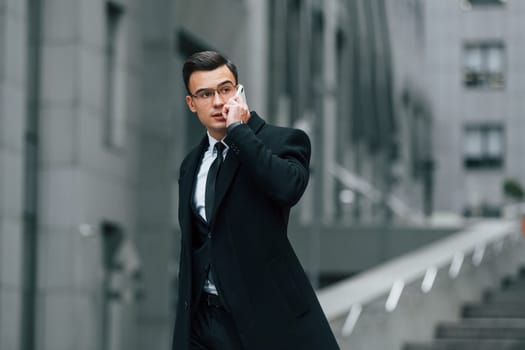 Talking by phone. Businessman in black suit and tie is outdoors in the city.