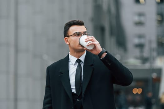 With cup of drink. Businessman in black suit and tie is outdoors in the city.