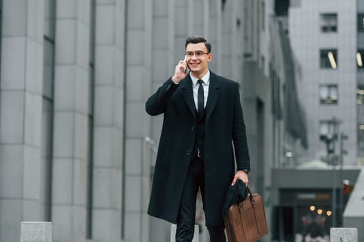 With smartphone. Businessman in black suit and tie is outdoors in the city.