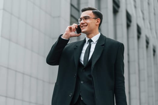 With smartphone. Businessman in black suit and tie is outdoors in the city.