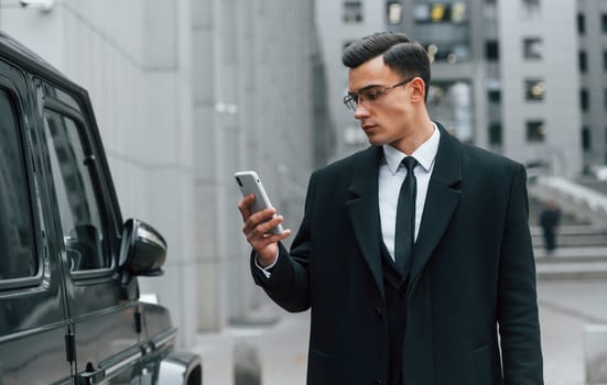Using phone. Businessman in black suit and tie is outdoors in the city.