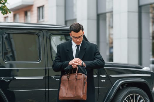 Holding brown bag in hands. Businessman in black suit and tie is outdoors in the city.