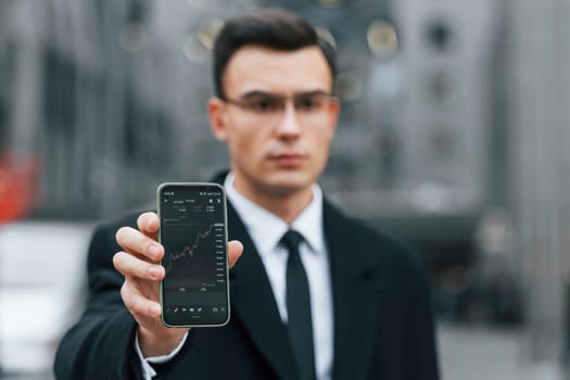 Holding smartphone with crypto graph in it. Businessman in black suit and tie is outdoors in the city.