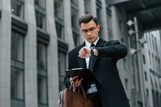 Looking at clock. Businessman in black suit and tie is outdoors in the city.