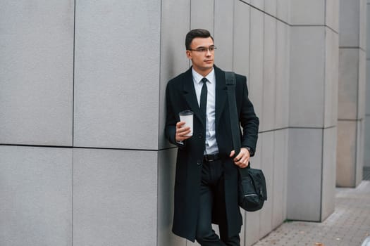 With cup of drink. Businessman in black suit and tie is outdoors in the city.