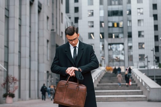 Looking at clock. Businessman in black suit and tie is outdoors in the city.