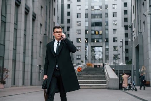 Talking by phone. Businessman in black suit and tie is outdoors in the city.