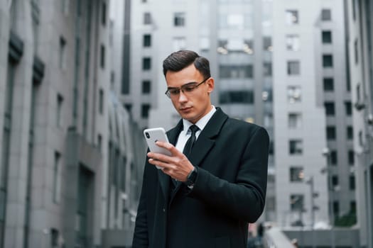 Phone in hand. Businessman in black suit and tie is outdoors in the city.