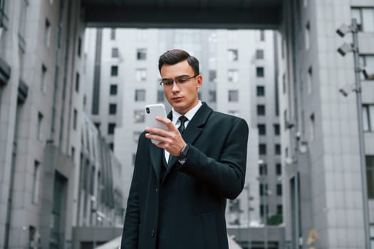 Phone in hand. Businessman in black suit and tie is outdoors in the city.