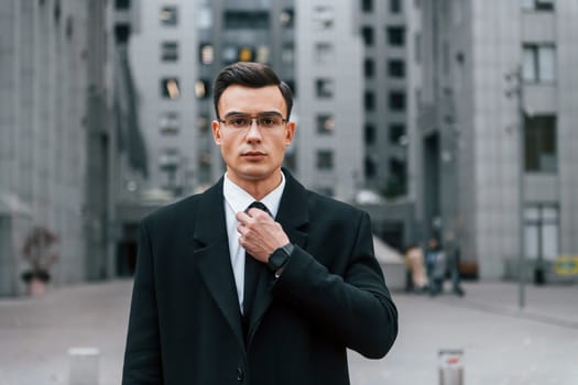 Fixing the tie. Businessman in black suit is outdoors in the city.