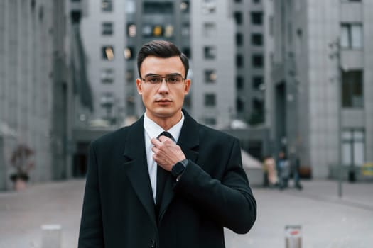 Fixing the tie. Businessman in black suit is outdoors in the city.