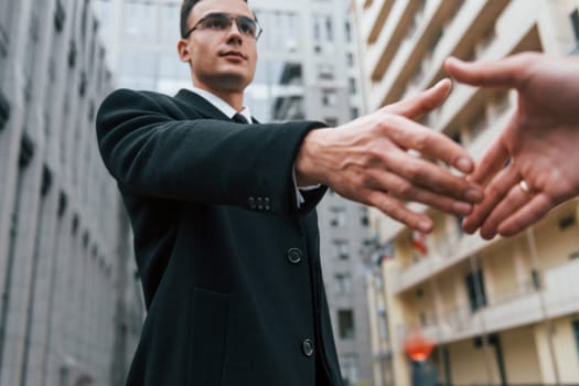 Doing handshake. Businessman in black suit and tie is outdoors in the city.