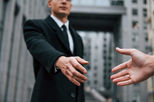 Doing handshake. Businessman in black suit and tie is outdoors in the city.