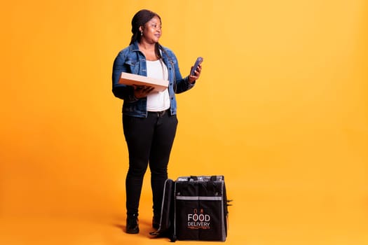 African american pizzeria courier holding pizza cardboard box while checking client adreess on smartphone before delivery order during lunch time. Food transportation service and takeaway concept