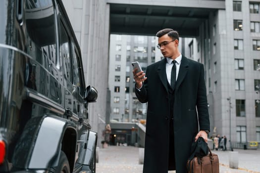 Standing near black car. Businessman in black suit and tie is outdoors in the city.