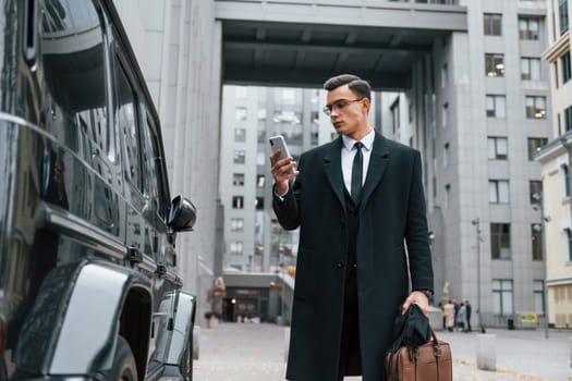 Standing near black car. Businessman in black suit and tie is outdoors in the city.