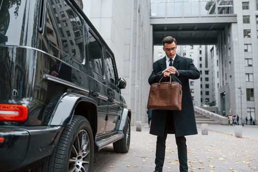 Standing near black car. Businessman in black suit and tie is outdoors in the city.