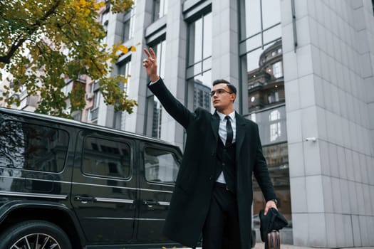 Standing against black car. Businessman in black suit and tie is outdoors in the city.