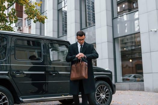 Standing against black car. Businessman in black suit and tie is outdoors in the city.