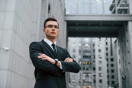 Standing with arms crossed. Businessman in black suit and tie is outdoors in the city.