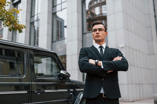 Standing with arms crossed. Businessman in black suit and tie is outdoors in the city.