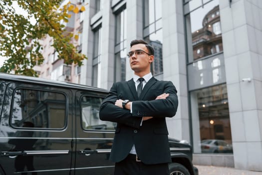 Standing with arms crossed. Businessman in black suit and tie is outdoors in the city.