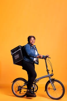 Pizzeria worker carrying food delivery thermal backpack standing riding bike in studio with yellow background. Restaurant courier delivering pizza for lunch. Food transportation service