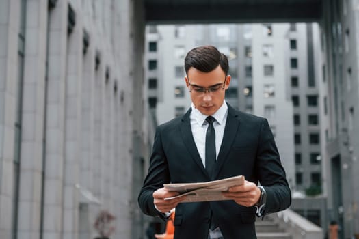 Reading some news. Businessman in black suit and tie is outdoors in the city.