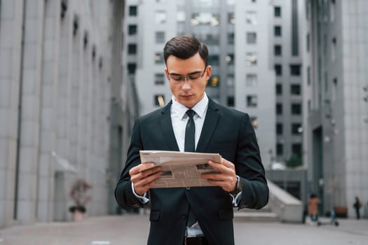Reading some news. Businessman in black suit and tie is outdoors in the city.