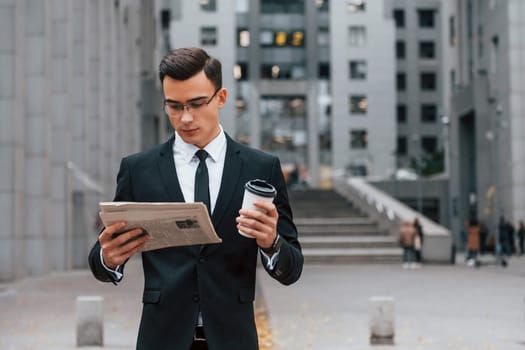 Reading some news. Businessman in black suit and tie is outdoors in the city.