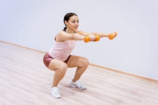 A woman of Caucasian ethnicity in sportswear squats with dumbbells in her hand against the background of a light wall. Home sports concept