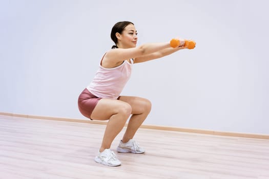 A woman of Caucasian ethnicity in sportswear squats with dumbbells in her hand against the background of a light wall. Home sports concept