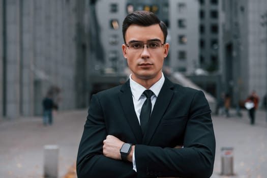 Standing with arms crossed. Businessman in black suit and tie is outdoors in the city.