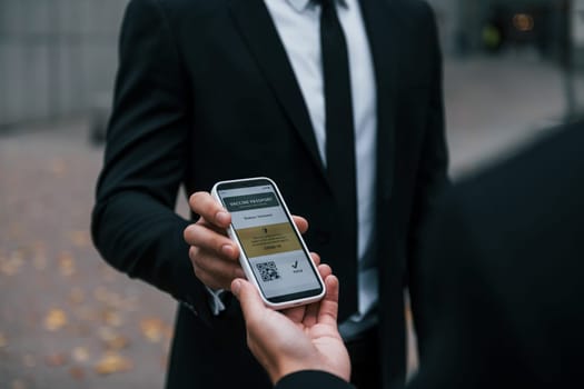 Holding phone with vaccination certificate. Businessman in black suit and tie is outdoors in the city.