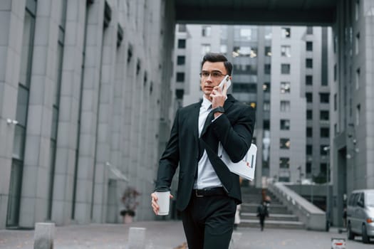 Walking and talking by phone. Businessman in black suit and tie is outdoors in the city.