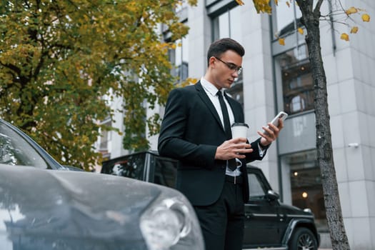 Near black car with phone in hands. Businessman in black suit and tie is outdoors in the city.