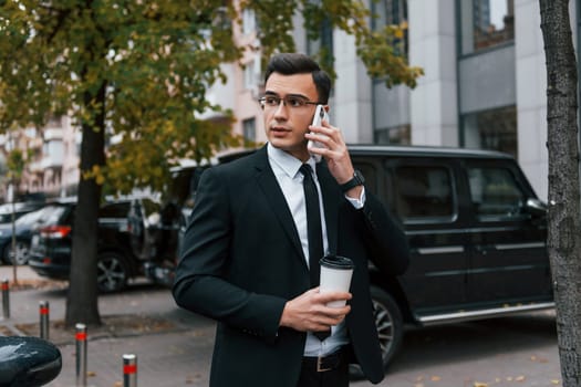 Near black car with phone in hands. Businessman in black suit and tie is outdoors in the city.