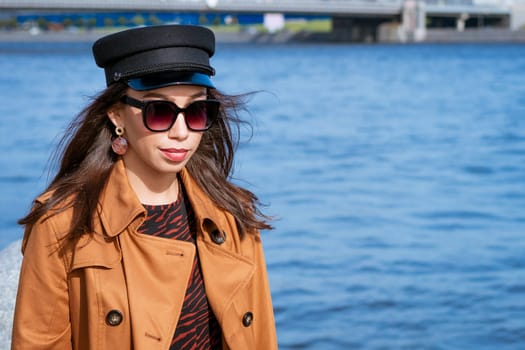 A female portrait of European appearance on the embankment on a sunny spring day with a newspaper in her hand. Beautiful brunette woman in sunglasses and in a brown jacket and black cap