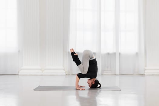 Side view of an athletic woman doing headstand training during yoga meditation in a bright gym. Concept of normalization of all processes in the body, strength, energy and concentration.