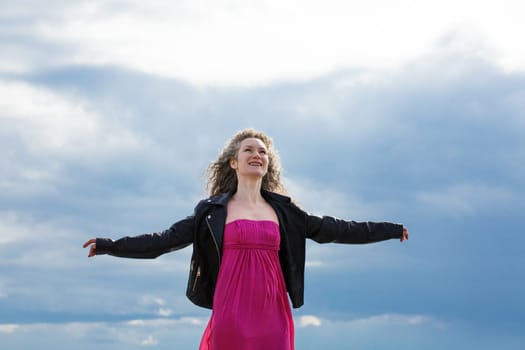 caucasian woman in a black leather jacket and a pink dress, spread her arms to the sides against the background of a gloomy sky