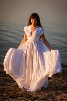 beautiful, young brunette woman in a white flowing dress on the sandy beach in the evening. full-length portrait in natural light.
