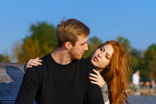 young couple of guy and girl with red hair of Caucasian appearance, in casual clothes, on a sunny day sitting in the park on a wooden bench in an embrace, happy relationship between a man and a woman