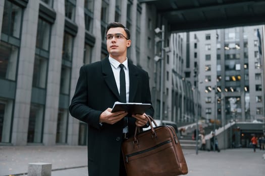 With notepad in hands. Businessman in black suit and tie is outdoors in the city.