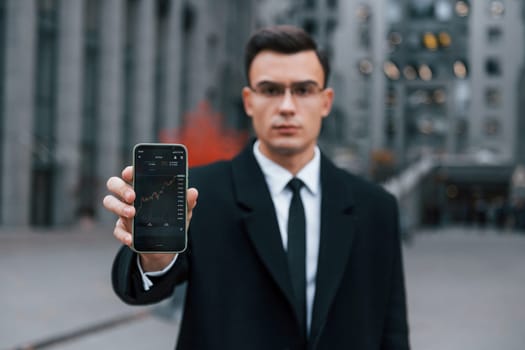 Showing phone with crypto graphs in it. Businessman in black suit and tie is outdoors in the city.