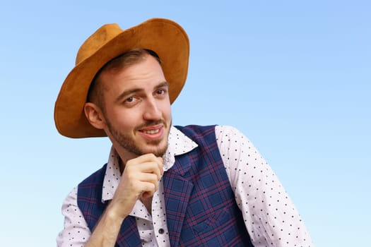 Portrait of a handsome young man in a hat against a blue sky. Caucasian guy with a goatee smiling happy summer outdoors sitting in a shirt and vest